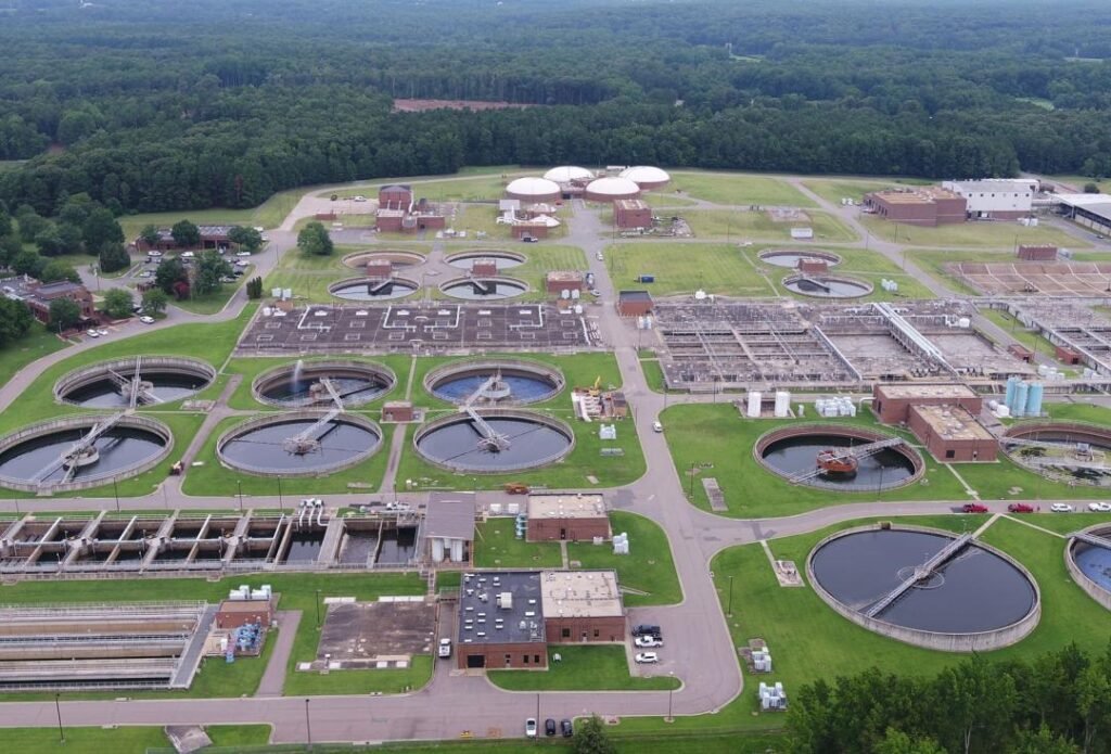 An aerial view of Lower Potomac Wastewater Treatment Plant showcases an expansive facility layout with multiple circular and rectangular tanks, pathways, and surrounding greenery. Several buildings and storage tanks are visible within this impressive plant.