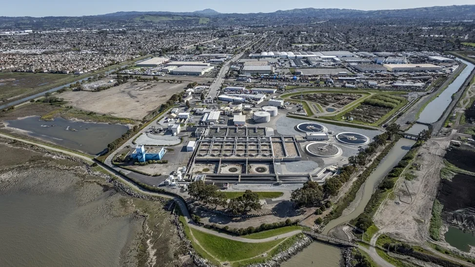 The aerial view showcases the T.E. Maxson Wastewater Treatment Plant near Memphis South, surrounded by water channels and lush greenery. In the background, a sprawling urban area stretches towards hills on the horizon under a clear sky.