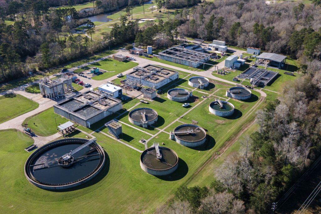 Aerial view of a wastewater treatment plant with circular and rectangular tanks surrounded by trees and grassy areas.