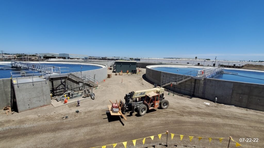 Wide-angle view of a construction site with two large circular water tanks at the Regional Wastewater Control Facility in Stockton. A JLG telehandler is parked in the dirt lot. Workers are visible near the tanks, and buildings can be seen in the distance under a clear blue sky.