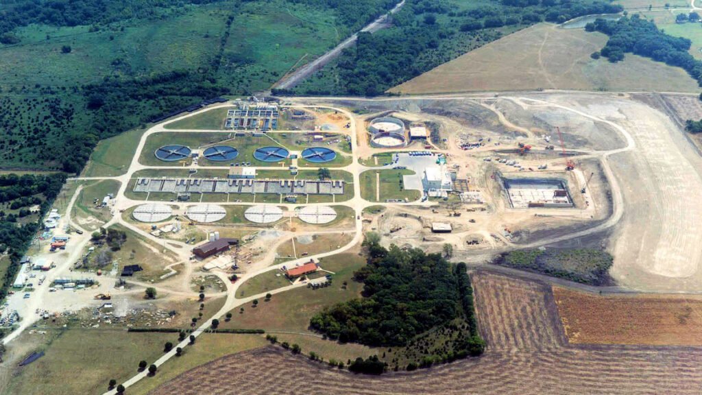 Aerial view of the Wilson Creek Regional Wastewater treatment plant surrounded by fields. The facility includes circular tanks, open basins, and various buildings. Roads connect different sections, with green areas and trees scattered around the site.