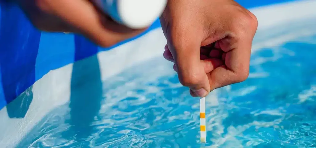 Hand testing pool water with a pH strip, showing color change for chemical balance in clear blue water.