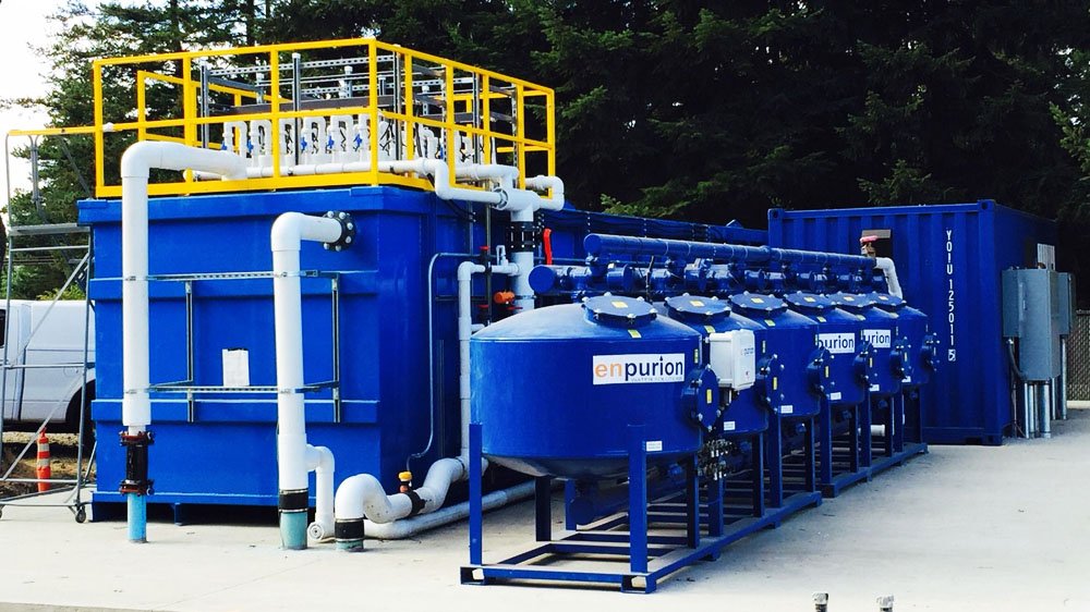 A blue industrial water treatment system equipped for electrocoagulation in wastewater features multiple tanks and connecting pipes. A yellow-railed platform tops the structure, surrounded by trees in the background and a paved surface in front.
