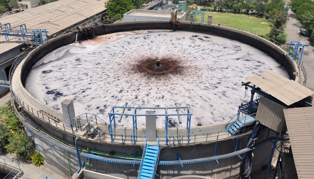 Aerial view of a large circular wastewater treatment tank with foamy liquid inside, surrounded by industrial structures.