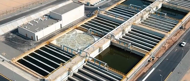 Aerial view of a wastewater treatment facility featuring a series of rectangular tanks and channels. These tanks, crucial for managing organic loading, are arrayed in an orderly grid pattern. Some contain dark water while others show varying stages of processing. Buildings and roads are nearby.