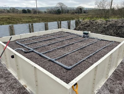 A rectangular elevated bed filled with gravel forms an integral part of the sand filter system for wastewater treatment. A network of pipes lies on top, surrounded by grass and a metal fence, with trees in the background under a cloudy sky. A shovel is propped against one side.