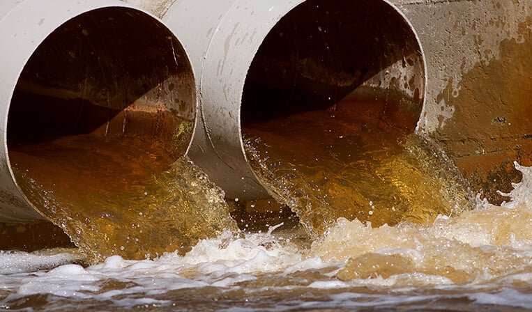 Two large pipes discharge brown wastewater into a body of water, causing foamy turbulence.