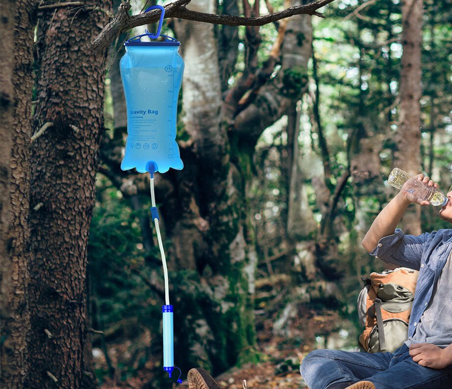 A blue gravity water filter bag hangs from a tree; a person sits nearby drinking from a bottle in a forest.