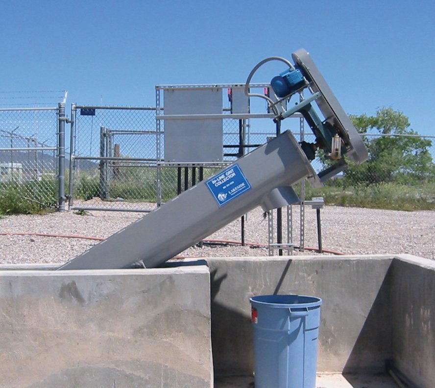 Industrial screw conveyor outdoors, angled with a blue motor and labeled "Spiral Grit Collector," near a blue trash bin.