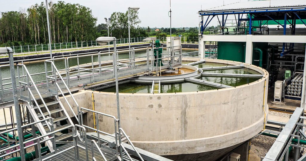 Industrial water treatment facility with large circular sedimentation tanks and metal walkways, surrounded by greenery.