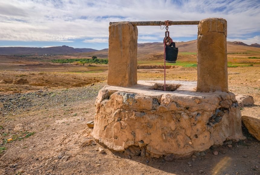 Ancient stone well with pulley and bucket in a dry landscape, under a partly cloudy sky.
