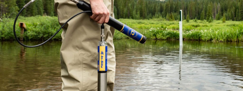 Person holding a water quality sensor in a river, surrounded by lush greenery and trees in the background.
