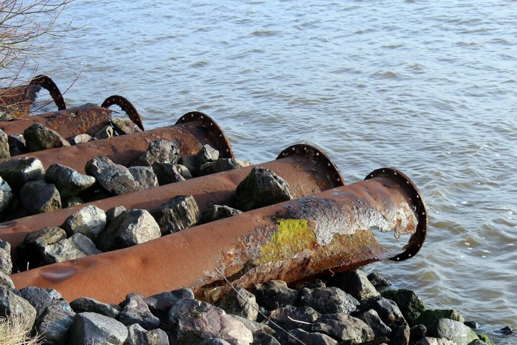 Rusty pipes extend into a body of water, surrounded by rocks.