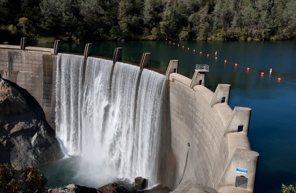 Concrete dam with cascading water, surrounded by lush trees and a calm lake.