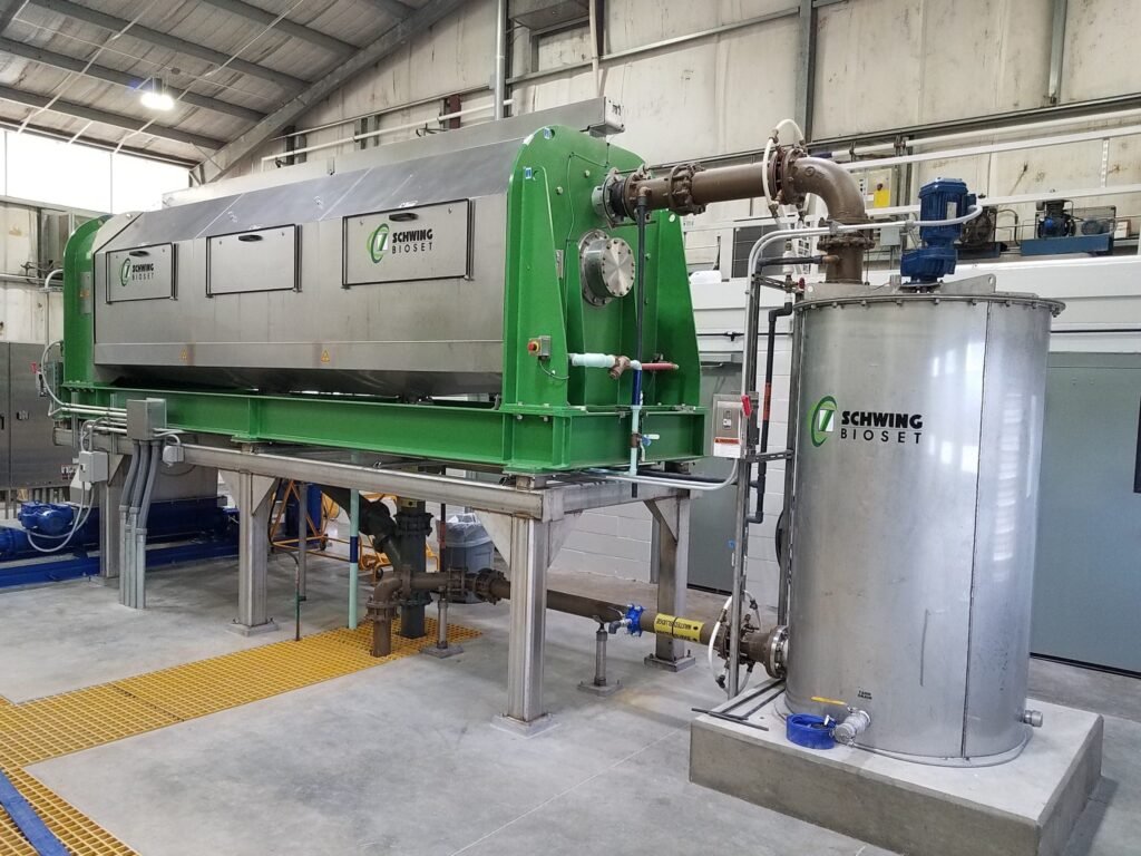 Industrial dewatering equipment by Schwing Bioset in a factory setting, featuring green and silver machinery indoors.