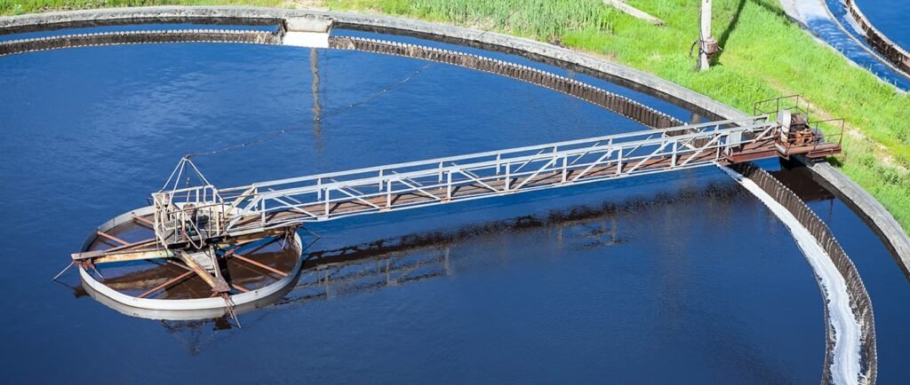 Aerial view of a circular wastewater treatment tank with a long metal bridge-like structure functioning as a skimmer across its diameter. The tank is filled with dark water, and green grass surrounds the area.