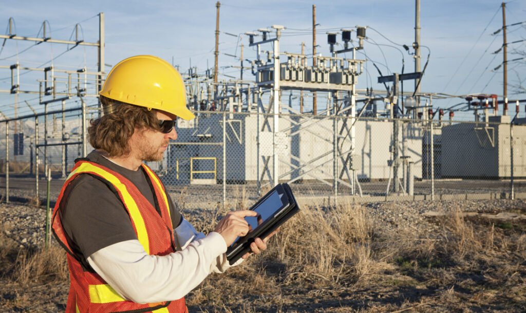 Worker in hard hat and safety vest using a tablet near electrical substation, with wires and equipment in background.