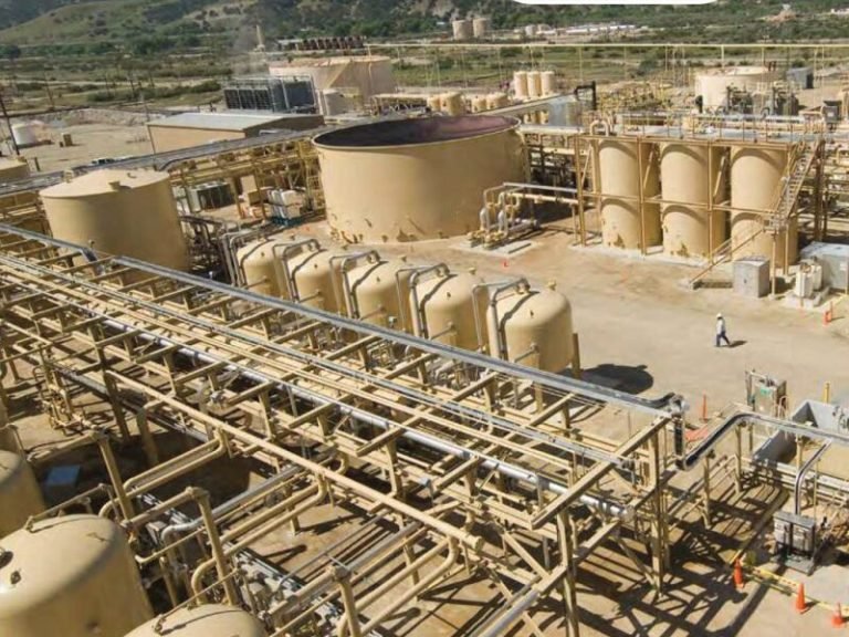 An industrial site showcases several large, cylindrical tanks and a network of pipes, tailored solutions for handling industry-specific applications. A person walks nearby, with a hilly landscape in the background. The scene unfolds from an elevated position.