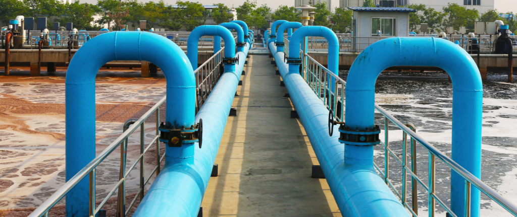 Blue pipes and railings line a walkway at a wastewater treatment facility with tanks of foamy water on either side.