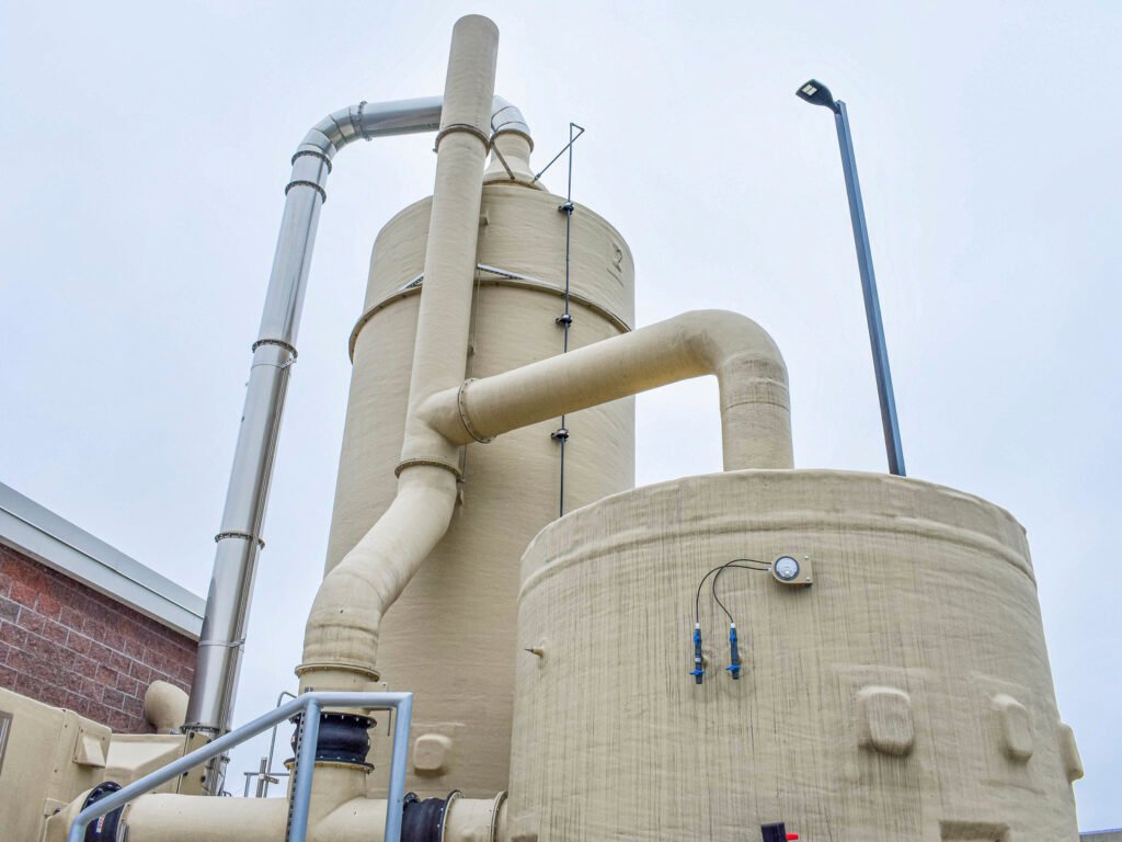 Industrial machinery with beige cylindrical tanks and pipes is shown against a cloudy sky, designed for efficient wastewater treatment. The setup includes a pressure gauge and mounted ladder, with metal piping connecting the structures. A brick wall is partially visible on the left.