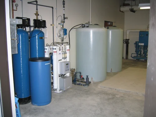 A room containing water treatment equipment, featuring ozone diffusers alongside blue and white tanks, pipes, and control panels. The gray concrete floor complements the beige walls, creating a harmonious industrial space.