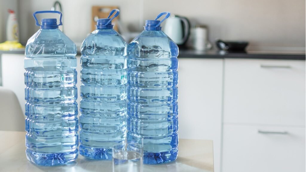 Three large blue plastic water bottles on a kitchen counter with a glass of water in the foreground.