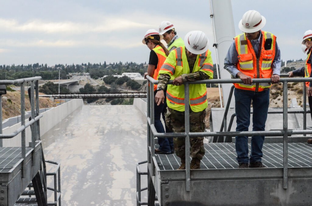 Workers in high-visibility gear and helmets inspect a large concrete structure on an overcast day.