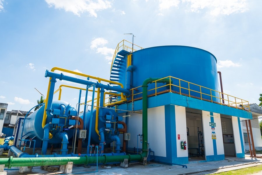 Blue industrial facility featuring large cylindrical tanks, pipes, and a ladder under a clear sky. The structure is part of an advanced treatment facility with a platform boasting yellow railings, surrounded by green and blue pipes, crucial for industrial wastewater management.