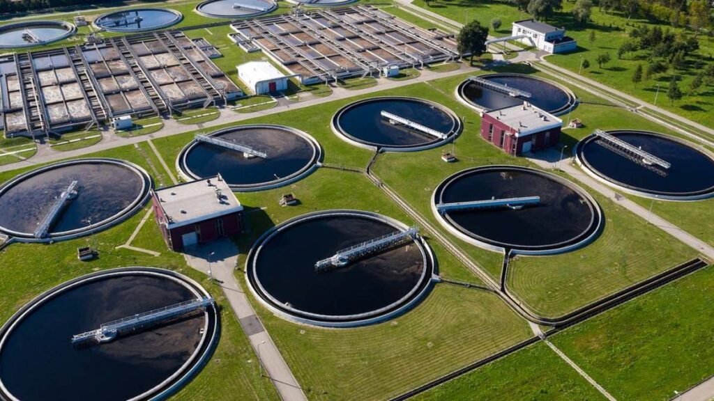 Aerial view of a cutting-edge wastewater treatment plant showcasing innovations with multiple large, circular tanks and pathways. The facility harmonizes urban water management, surrounded by lush green grass and trees, with several buildings interspersed among the tanks.